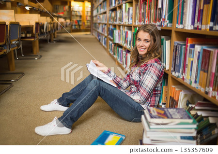 Female student sitting on carpeted library aisle reading book with textbooks and notebook nearby Female student sitting on carpeted library aisle reading book with textbooks and notebook nearby 135357609