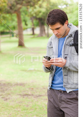 Man in early twenties standing in urban park checking smartphone and carrying backpack, copy space 135357620