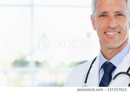 Senior male doctor standing in office by window wearing lab coat holding stethoscope, copy space 135357622