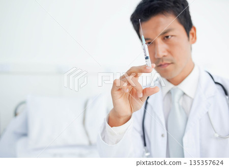 Asian male doctor inspecting syringe needle with stethoscope on neck at clinic bed, copy space 135357624