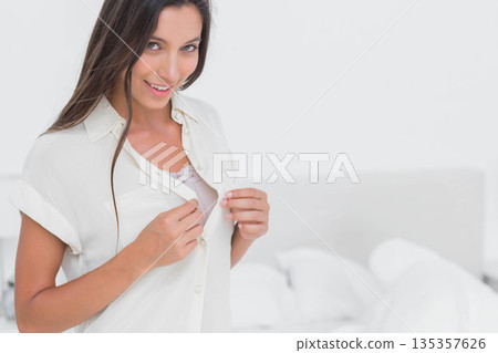 Woman holding white button-up shirt revealing lace-trimmed camisole in bright bedroom, copy space Woman holding white button-up shirt revealing lace-trimmed camisole in bright bedroom, copy space 135357626