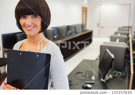 Female instructor standing left in computer lab holding clipboard near monitors, copy space 135357630