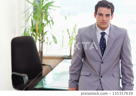 Male professional leaning against glass-top desk in office wearing suit and tie near potted plant Male professional leaning against glass-top desk in office wearing suit and tie near potted plant 135357639