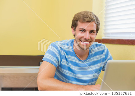 Male student sitting at wooden desk in study nook working on silver laptop beside windowsill blinds 135357651