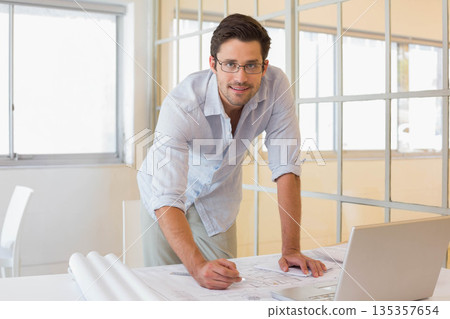 Male professional leaning over table in office drawing blueprints with pencil next to open laptop Male professional leaning over table in office drawing blueprints with pencil next to open laptop 135357654