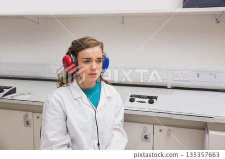 Woman in twenties sitting in exam room wearing lab coat, red and blue headphones using audiometer Woman in twenties sitting in exam room wearing lab coat, red and blue headphones using audiometer 135357663