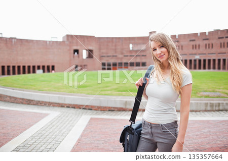 Female student posing with messenger bag beside concrete border with brick facade, copy space 135357664