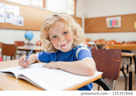 Blond boy leaning on one arm writing in open workbook at wooden school desk with pencil 135357666
