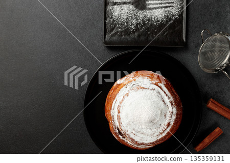 Round, delicious bun with cinnamon and powdered sugar on a black plate on a gray background Round, delicious bun with cinnamon and powdered sugar on a black plate on a gray background 135359131