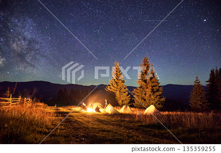 Blurred silhouettes of hikers resting around campfire, surrounded by illuminated tents under stunning starry sky. Milky Way stretches above, creating magical atmosphere in mountain wilderness. Blurred silhouettes of hikers resting around campfire, surrounded by illuminated tents under stunning starry sky. Milky Way stretches above, creating magical atmosphere in mountain wilderness. 135359255