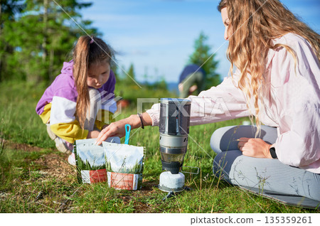 Tourist woman and young girl preparing meal from packaged pouch using portable camping stove on grassy hillside. Scenic mountain backdrop adds to serene outdoor setting. Tourist woman and young girl preparing meal from packaged pouch using portable camping stove on grassy hillside. Scenic mountain backdrop adds to serene outdoor setting. 135359261