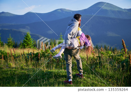 Father joyfully playing with his daughter, as they share playful moment in meadow. Both laughing, with backdrop of lush green hills and distant mountains under clear blue sky. Father joyfully playing with his daughter, as they share playful moment in meadow. Both laughing, with backdrop of lush green hills and distant mountains under clear blue sky. 135359264