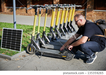 Man connects solar panel to electric scooter for charge, highlighting practical application of sustainable renewable energy. Concept of integration of eco-friendly technology in urban transportation. Man connects solar panel to electric scooter for charge, highlighting practical application of sustainable renewable energy. Concept of integration of eco-friendly technology in urban transportation. 135359265
