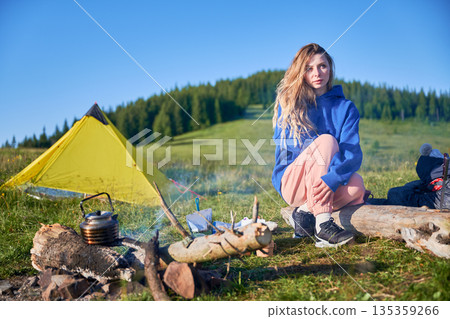 Young woman in resting on log next to campfire, looking thoughtful. Yellow tent and camping gear nearby. Lush, green mountain meadow under clear blue sky, with forested hill in background. Young woman in resting on log next to campfire, looking thoughtful. Yellow tent and camping gear nearby. Lush, green mountain meadow under clear blue sky, with forested hill in background. 135359266
