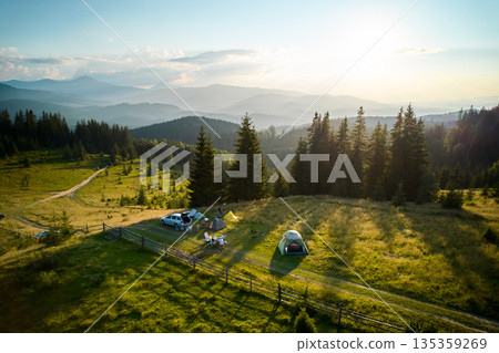 Aerial view of picturesque campsite on grassy hill, surrounded by evergreen trees and overlooking mountain vista. Distant mountains, tourist tents, cars, camping gear under golden evening light. 135359269