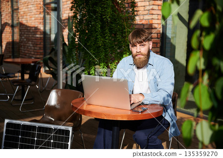 Man works on laptop and smartphone at outdoor table. Solar panel nearby, emphasizing sustainable, modern eco-friendly workspace that combines technology and sustainability. Man works on laptop and smartphone at outdoor table. Solar panel nearby, emphasizing sustainable, modern eco-friendly workspace that combines technology and sustainability. 135359270
