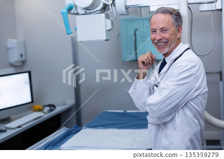 Senior male doctor standing by X-ray machine in radiology room wearing lab coat, stethoscope Senior male doctor standing by X-ray machine in radiology room wearing lab coat, stethoscope 135359279