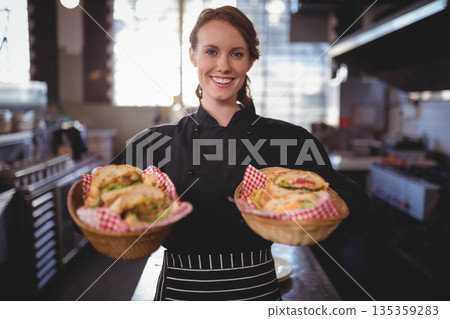 Female chef in uniform holding wicker baskets with checkered liners, sandwiches, fries in kitchen 135359283