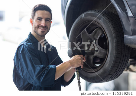 Male mechanic standing by raised car wheel removing lug nuts with pneumatic wrench in garage 135359291