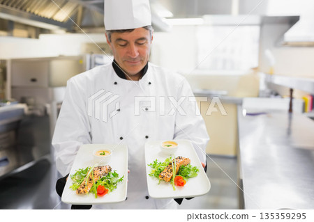 Senior male chef wearing chef coat presenting two seared fish plates with sauce, greens in kitchen 135359295