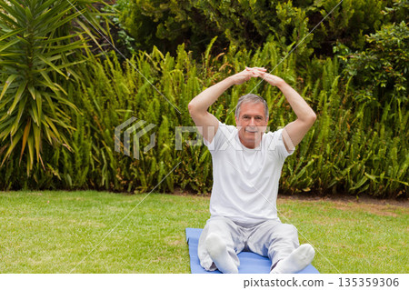 Senior man wearing exercise clothes stretching both arms overhead on blue exercise mat in backyard Senior man wearing exercise clothes stretching both arms overhead on blue exercise mat in backyard 135359306