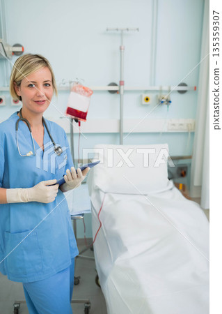 Female nurse holding touchscreen tablet in medical bay beside bed with IV bag, copy space 135359307