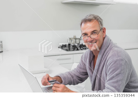Senior man wearing bathrobe holding credit card, typing on laptop at kitchen island, copy space Senior man wearing bathrobe holding credit card, typing on laptop at kitchen island, copy space 135359344