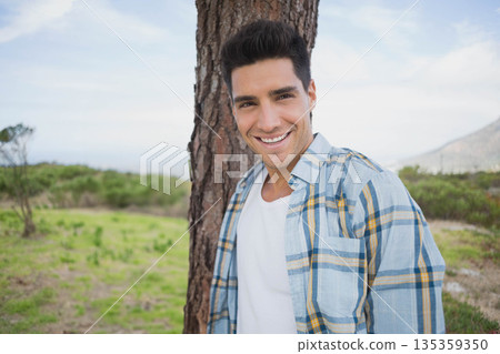 Man in his twenties leaning against tree trunk in grassy clearing with rolling hills Man in his twenties leaning against tree trunk in grassy clearing with rolling hills 135359350