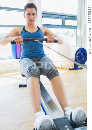 Woman pulling rowing handle on machine in fitness studio wearing blue tank top and gray leggings Woman pulling rowing handle on machine in fitness studio wearing blue tank top and gray leggings 135359354