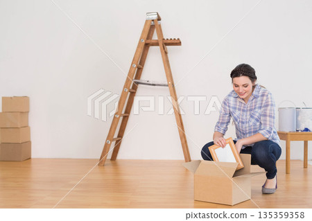 Woman kneeling and unpacking frame from box in empty room with stepladder, paint cans, copy space Woman kneeling and unpacking frame from box in empty room with stepladder, paint cans, copy space 135359358