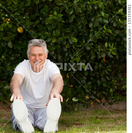 Senior man stretching forward toward toes on grass in backyard garden wearing white athletic shoes 135359361