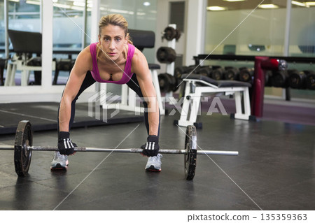 Female athlete bending over and gripping barbell in gym wearing pink tank top, black leggings Female athlete bending over and gripping barbell in gym wearing pink tank top, black leggings 135359363