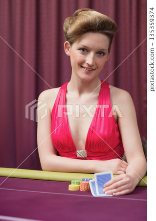 Female poker player in red dress sitting at casino poker table holding cards over chip stacks 135359374
