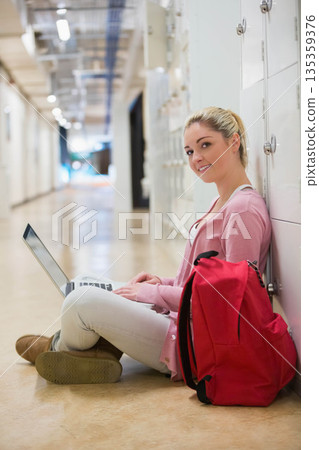 Female student sitting in hallway leaning on lockers using laptop beside red backpack, copy space 135359376