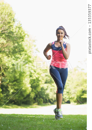 Female runner jogging on lawn in sunlit public park wearing navy cropped leggings and running shoes 135359377
