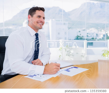 Man in shirt and tie sitting at office desk reviewing printed charts with pen and notebook Man in shirt and tie sitting at office desk reviewing printed charts with pen and notebook 135359380
