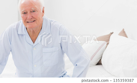 Senior man sitting on bed wearing blue striped pajamas and smiling in bedroom with white linens 135359387