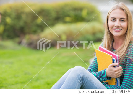 Smiling woman sitting on grass wearing striped shirt holding colorful notebooks at park, copy space 135359389