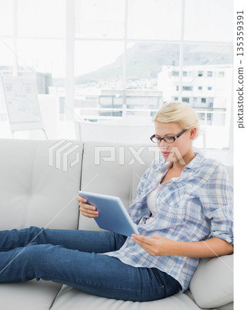 woman wearing glasses reading tablet on leather couch beside whiteboard in living room, copy space woman wearing glasses reading tablet on leather couch beside whiteboard in living room, copy space 135359391
