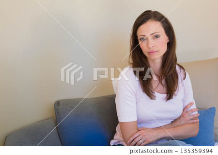 Middle-aged woman sitting with arms crossed on grey couch with blue and neutral pillows, copy space Middle-aged woman sitting with arms crossed on grey couch with blue and neutral pillows, copy space 135359394