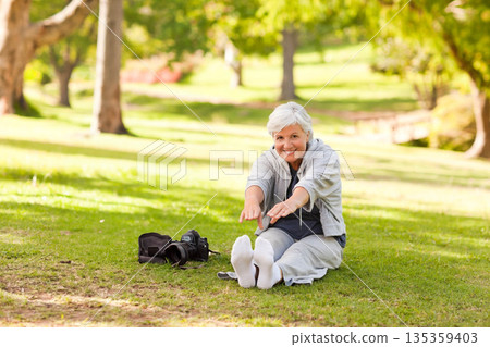 Senior woman wearing tracksuit stretching on grass in park with camera and black camera bag Senior woman wearing tracksuit stretching on grass in park with camera and black camera bag 135359403