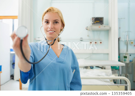 Female nurse holding stethoscope toward camera in hospital exam room with bed and medical equipment 135359405