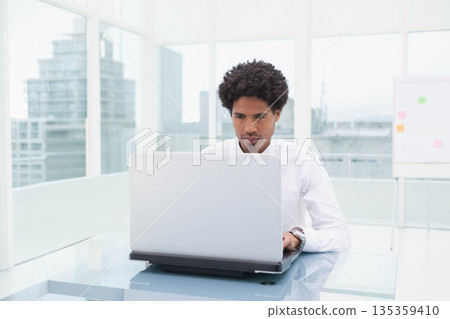 African American man typing on silver laptop at glass desk near whiteboard in office 135359410