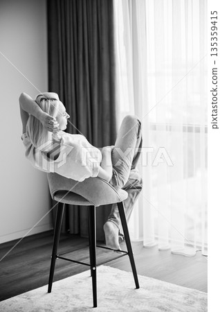 Back view of gorgeous woman relaxing on bar stool. Pretty female sitting on stool with her legs crossed. Woman in blue shirt and jeans looking in window. Black and white image. Back view of gorgeous woman relaxing on bar stool. Pretty female sitting on stool with her legs crossed. Woman in blue shirt and jeans looking in window. Black and white image. 135359415