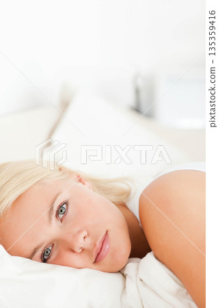 Woman lying on bed in bright bedroom wearing white tank top with white pillow and sheets Woman lying on bed in bright bedroom wearing white tank top with white pillow and sheets 135359416