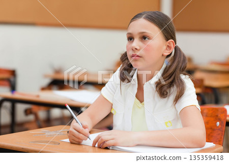 Female child holding pencil while writing in open notebook at school desk in classroom, copy space Female child holding pencil while writing in open notebook at school desk in classroom, copy space 135359418