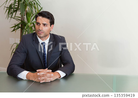 Man sitting at glass-topped conference table, clasping hands beside potted plant in suit and tie Man sitting at glass-topped conference table, clasping hands beside potted plant in suit and tie 135359419