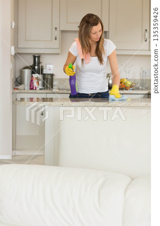 Woman wearing gloves cleaning counter in home kitchen, wiping granite with purple spray and cloth 135359426