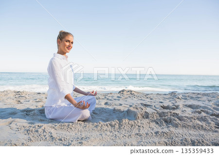 Woman meditating cross-legged on sandy shoreline facing calm sea wearing white clothing, copy space 135359433