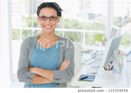 Woman standing at office window viewing trees, smiling wearing glasses, showing desk tech and plant 135359436